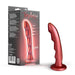 A Jealousy 7 Inch Rippled Silicone Dildo in red, made by Blush, is shown alongside its packaging that highlights the brand and product information. The dildo features a unique, wave-like design and textured surface with a flared base to enhance g-spot stimulation for varied sensations.