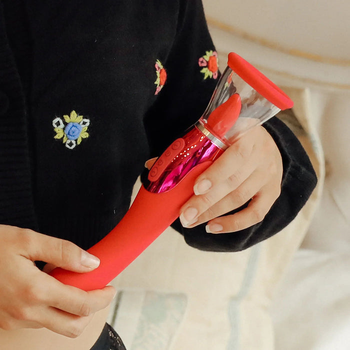 A close-up of a person holding the pink Jimmyjane Apex Double Ended Licking, Sucking G-Spot Vibrator by Pipedream Products. The vibrator features a transparent top and control buttons in the middle. The person is wearing a black sweater with colorful floral embroidery, and they are indoors with a blurred background.