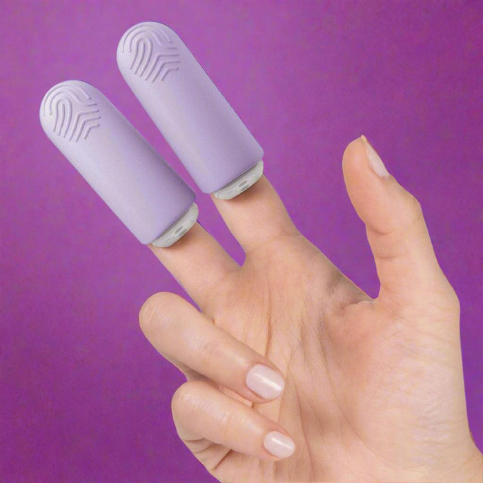 A hand with neatly manicured nails has two fingers fitted with flat, purple silicone finger sleeves from the Jimmyjane Hello Touch Pro Two Bullets set by Pipedream Products. The protectors feature textured, wave-like patterns on the tips. The background is white.