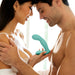 A woman and a man, both minimally clothed, stand close together with happy expressions, holding a Jimmyjane Pulsus Hands-Free G-Spot Fingering Vibrator with Remote in teal between them. The softly lit background suggests an intimate and tender moment.