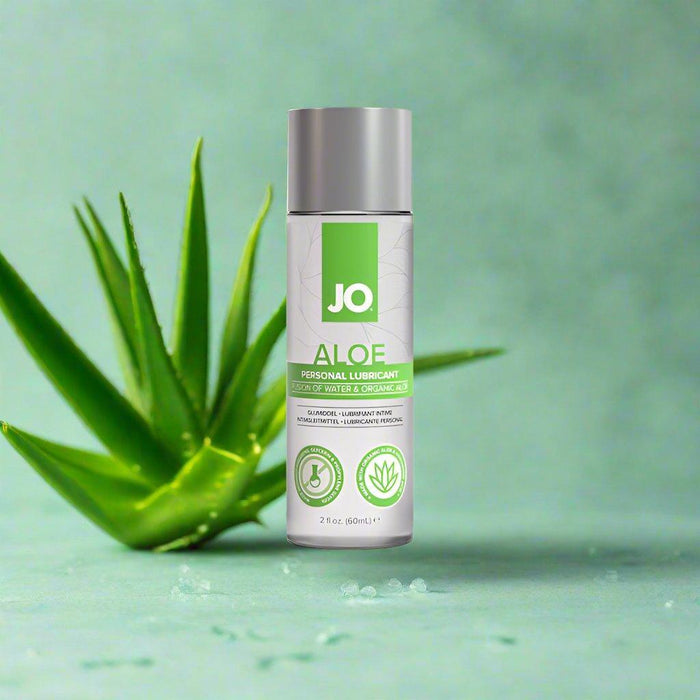 A small bottle of Jo Aloe Water Based Sensitive Skin Lubricant from System Jo stands upright on a light green surface, with a green aloe plant in the background. The lubricant bottle, featuring a metallic cap and an organic aloe label, boasts product details that highlight its pH-balanced formula for optimal comfort.