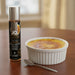 A 1oz bottle of Jo Gelato Creme Brulee Flavored Lubricant sits beside a white ramekin of crème brûlée and spoon on a wooden table, highlighting this indulgent dessert-inspired lube with a softly blurred background.