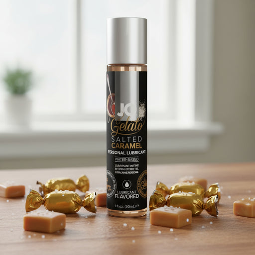 A 1oz bottle of Jo Gelato Salted Caramel Flavored Lubricant sits on a wooden surface, surrounded by caramel candies and sprinkled salt, with a blurred window in the background.