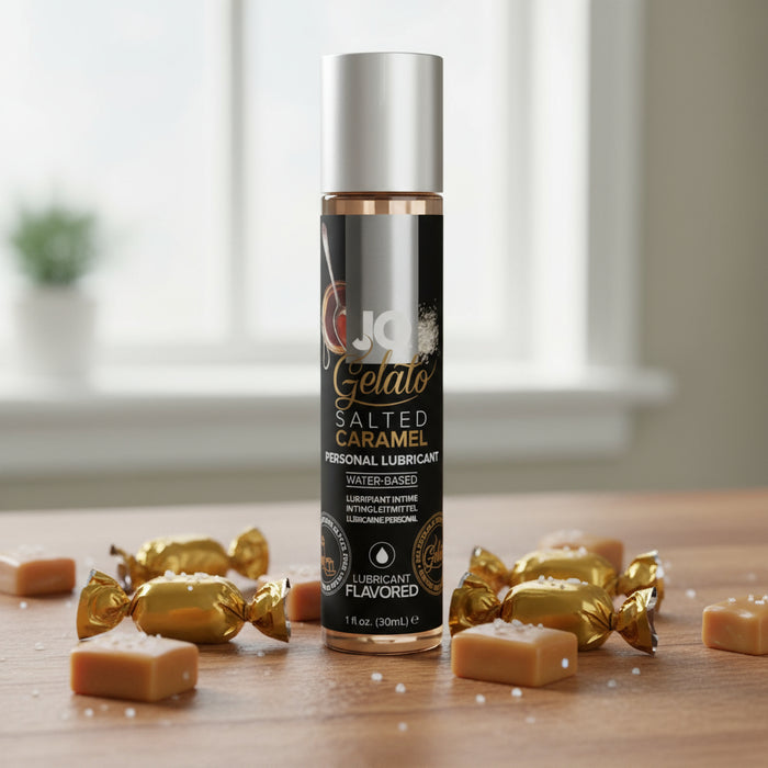 A 1oz bottle of Jo Gelato Salted Caramel Flavored Lubricant sits on a wooden surface, surrounded by caramel candies and sprinkled salt, with a blurred window in the background.