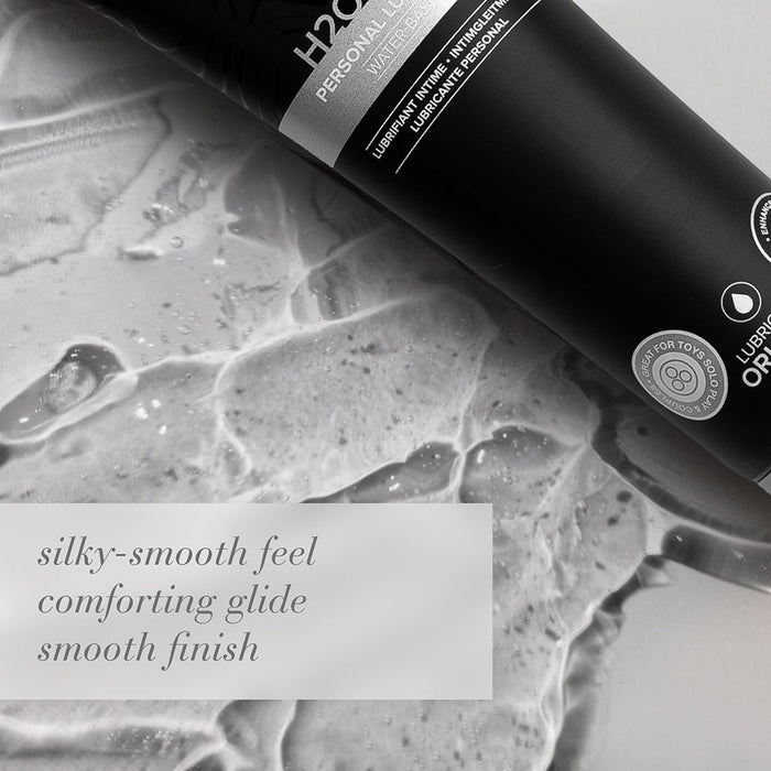 A black tube of JO H2O Gel Ultra Thick Water Based Lubricant from System Jo rests on a wet, glossy surface. Next to it, in light font, reads: "silky-smooth feel comforting glide smooth finish." The packaging is adorned with a circle logo and detailed text highlighting that the product is moisturizing and odor-free.