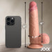 A smartphone rests beside the Jock Real Skin 8 Inch Silicone Suction Cup Dildo with Balls in a caramel shade. This realistic item from XR Brands features a super strong suction cup, measuring 8 inches in height, with a circumference of 6 inches and a diameter of 1.7 inches. The name "Jock" is delicately displayed in the corner, highlighting its dual density design for an authentic experience.