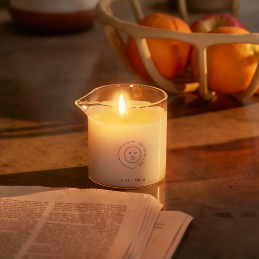 The Wild Lust Massage Oil Candle by Dame (Lime Leaf, Ambrette & White Peach) in a glass beaker rests on a table beside an open newspaper, with a bowl of oranges behind it. Warm, natural light enhances the cozy ambience.