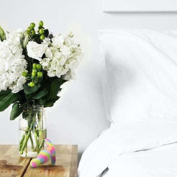 A clear glass vase with white hydrangeas and green berries sits on a wooden bedside table next to a white pillow and white blanket. Beside the vase is the Blush Kaleido Silicone Beginner's Butt Plug, featuring a brightly-colored pink and blue cone shape made from easy-to-clean platinum-cured silicone.