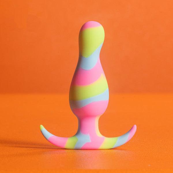 The Kaleido Silicone Beginner's Butt Plug by Blush is a colorful, platinum-cured silicone toy adorned with a spiraled pattern of pink, green, and blue hues. It features a flared base with two crescent-shaped extensions and stands upright on an orange surface against an orange background—perfect for anal beginners and easy to clean.