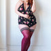 A woman with red hair is posing confidently against a white wall. She is wearing a floral print lingerie set with black lace trim and dark red **Kix'ies Heather Cabernet Opaque Thigh Highs (sizes A-D)** with no-slip-grip and black stripes at the top. She has bright red lipstick on and is looking off to the side.