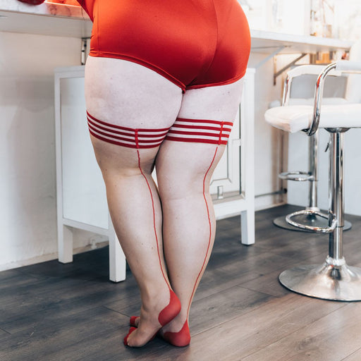 A person wearing a red bodysuit and Kix'ies Whitney Red Back Seam Sheer Thigh Highs (sizes A-D) with horizontal stripes and no-slip-grip stands on a wooden floor. They are facing a countertop, their back to the camera. A white bar stool is visible on the right.