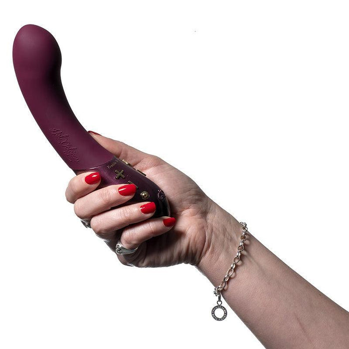 A hand with red painted nails and a silver bracelet holds the maroon Kurve Dual Motor G-Spot Vibrator by Hot Octopuss, featuring control buttons near the base, against a white background.