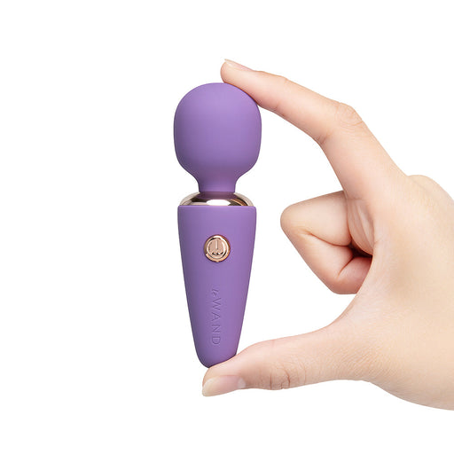 A hand holds the Le Wand Micro Powerful Pocket Sized Wand, a small purple mini massager with a rounded top and gold button, isolated on white. This travel-friendly vibrator offers convenience wherever you go.