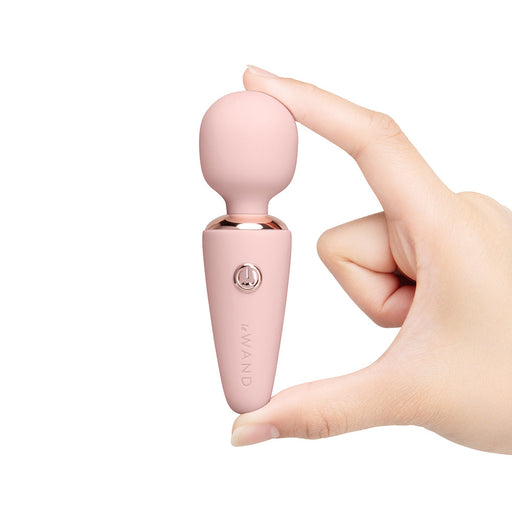 A hand holds the Le Wand Micro Powerful Pocket Sized Wand, a small pink mini massager with a rounded top and power button, known for deep rumbly vibrations, against a white background.