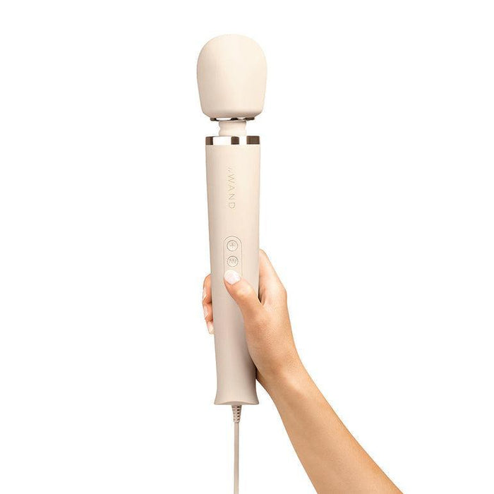 A hand holding a cream-colored Le Wand Plug-In Powerful Wand Vibrator with a rounded head. This Le Wand device features two buttons on its handle, offering 10 vibration levels for customizable rumbly vibes, and is attached to a cord.