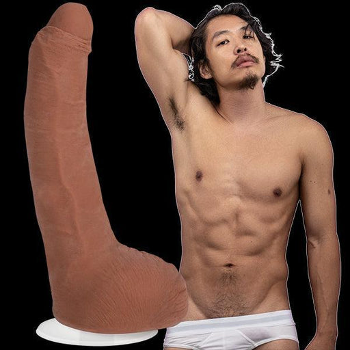 A shirtless man with medium-length dark hair and a mustache poses confidently with his right arm resting behind his head. He is wearing white underwear. To his left, there is a large, realistic Leo Vice Uncut Vac U Lock Porn Star Dildo - Caramel by Doc Johnson, made from Ultraskyn material and mounted on a white base.