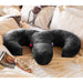 A black Liberator Arie Toy Mount Snuggle Pillow with two vibrator pockets lies on an unmade bed with white sheets and a bra; nearby are a magazine, wireless headphones, and a small pink item partly under the pillow.