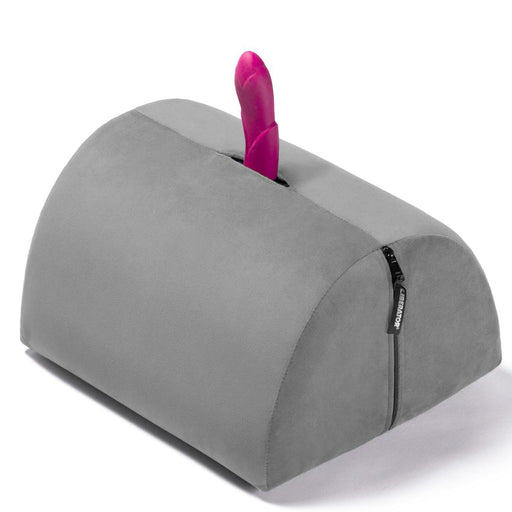 The Liberator BonBon Rideable Sex Toy Mount is a gray, semi-cylindrical foam cushion with a zippered side and a top slot designed to hold a pink silicone dildo upright for sturdy, hands-free play.