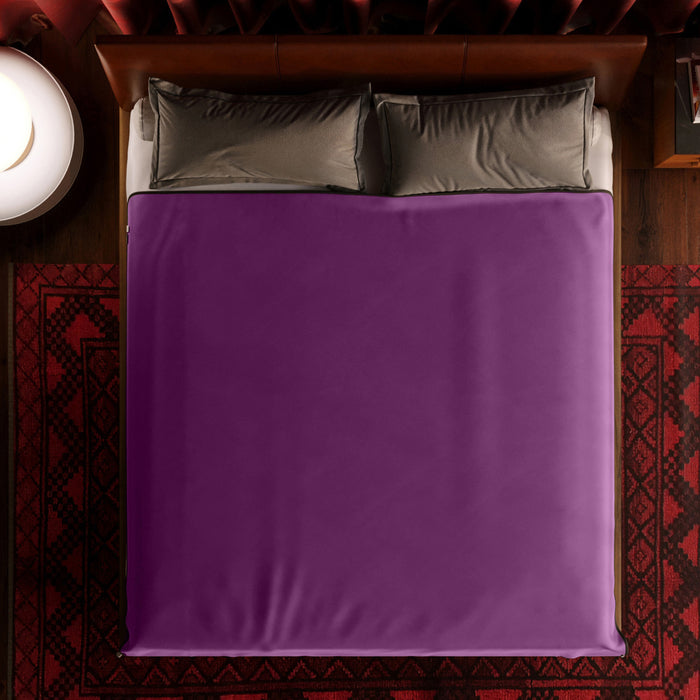 A neatly made bed with gray pillows features the Liberator Fascinator Throw King Size Waterproof Sex Blanket in purple, viewed from above. The bed sits on a patterned red rug, with a round white lamp on the left side.