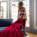 A woman in black lingerie and stockings stands by a door, holding a red velvet robe that drapes over a couch and onto the Liberator Fascinator Throw King Size Waterproof Sex Blanket spread across the wooden floor. Sunlight pours through glass doors behind her.