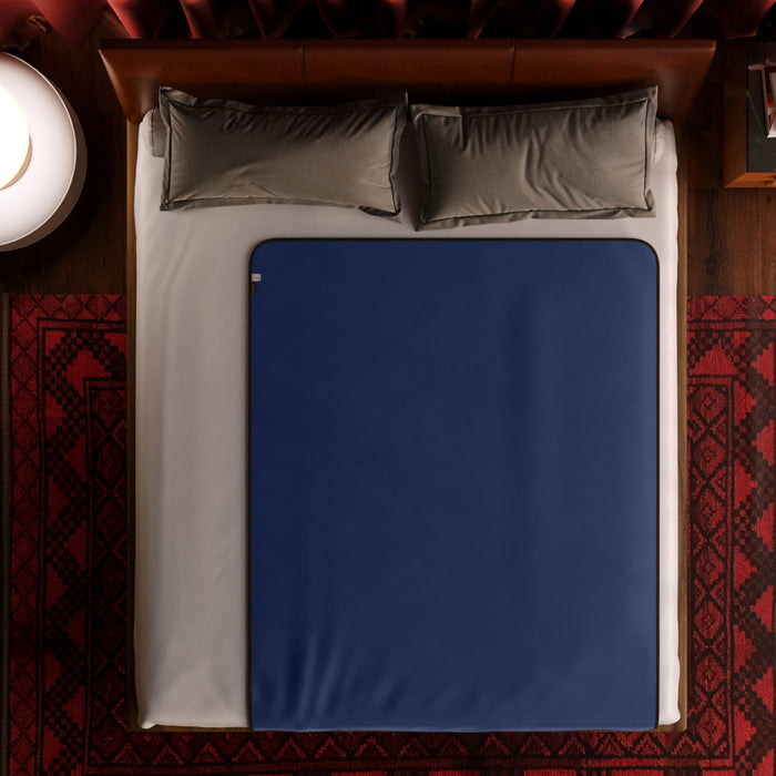 A neatly made bed with two gray pillows and a dark blue Liberator Fascinator Throw Queen Size Waterproof Sex Blanket over white sheets, surrounded by a red patterned rug and featuring a side table with a round white lamp.