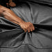 A close-up shows a hand gripping and pulling at the Liberator Fascinator Throw Queen Size Waterproof Sex Blanket in dark gray, forming deep folds in the fabric. A tensed arm is partially visible.