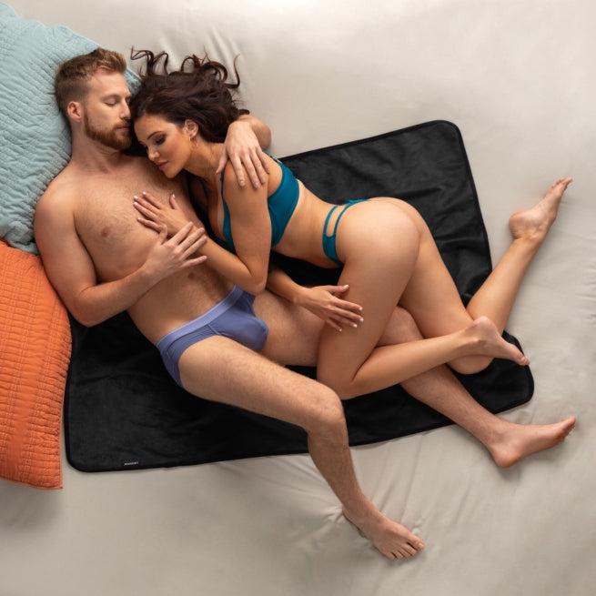 A man and woman in underwear cuddle closely on a bed atop the Liberator Fascinator Travel Sized Waterproof Sex Blanket, with colorful pillows nearby.