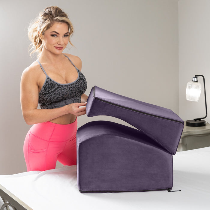 A woman in a gray sports bra and bright pink leggings places the Liberator Flip Ramp Convertible Sex Positioning Furniture on another cushion on a bed, with a lamp and discreet ottoman visible in the background.