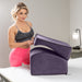 A woman in a gray sports bra and bright pink leggings places the Liberator Flip Ramp Convertible Sex Positioning Furniture on another cushion on a bed, with a lamp and discreet ottoman visible in the background.