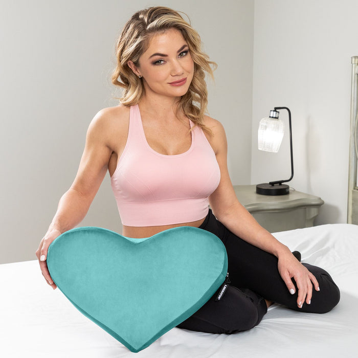 A woman in a pink sports bra sits on a bed, smiling and holding the Liberator Heart Wedge Sex Positioning Cushion, which is large and teal. A lamp and nightstand are visible in the background.
