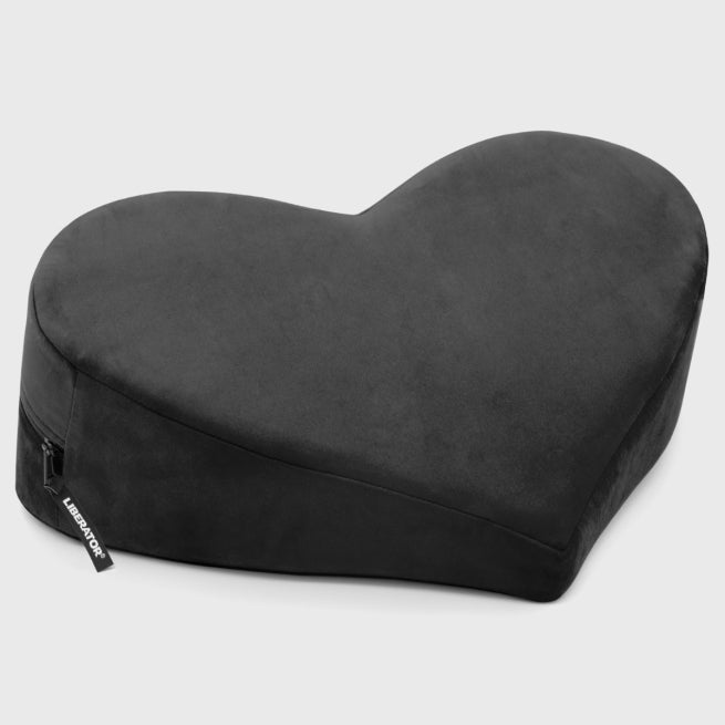 The Liberator Heart Wedge Sex Positioning Cushion, made of black wedge foam with a soft fabric cover and a side tag bearing the brand name, is displayed on a light gray background.