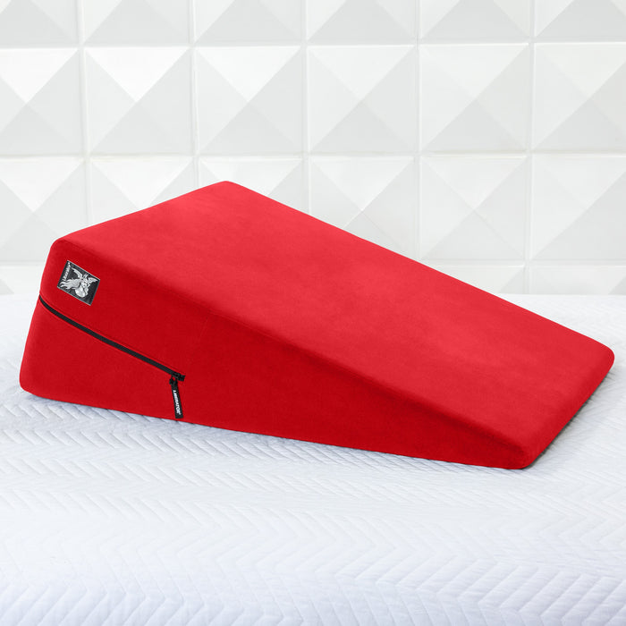 A vibrant red Liberator Ramp Sex Positioning Cushion - 30 Inches with a zippered cover rests on a white textured bedspread, set against a geometric white wall.