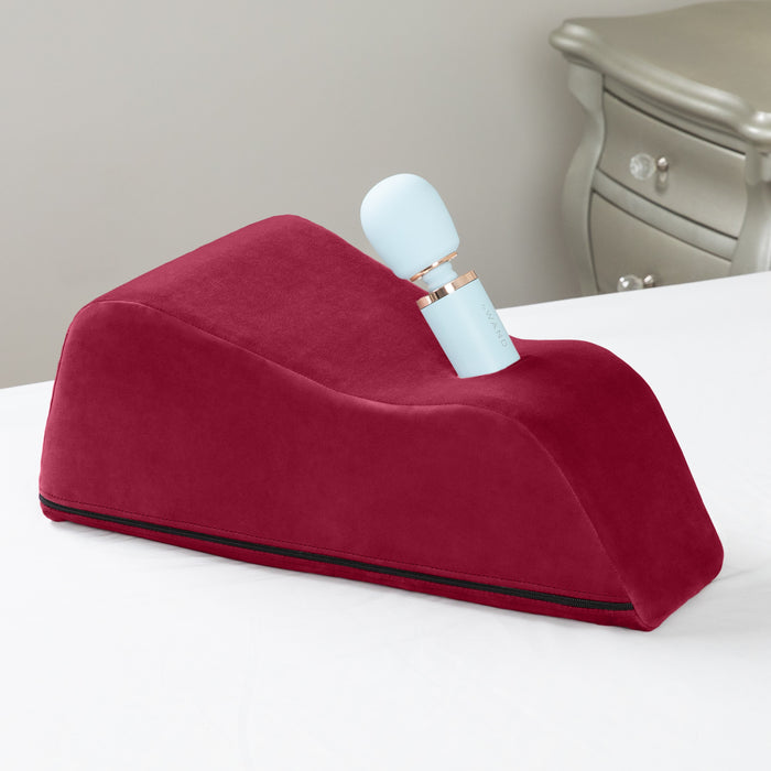 The Liberator Wanda – Hands-Free Wand Mount is a red, wedge-shaped pillow with an angled groove holding a light blue Magic Wand vibrator, shown on a white surface with a silver nightstand in the background.