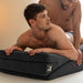 Two shirtless men are on a bed using the Liberator Wedge + Ramp Combo – The Iconic Duo for Deeper Pleasure. One lies on his stomach atop the support while the other leans over him from behind, in a softly lit room with neutral walls.