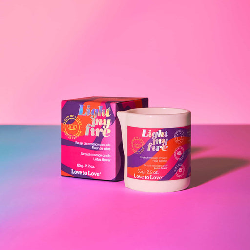 The Light My Fire Lotus Flower Scent Sensual Massage Candle sits beside its colorful box, both featuring bold patterns and bringing sensual vibes to your space on a pink and purple gradient surface.