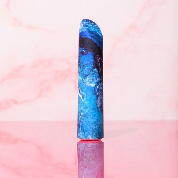 A cylindrical object featuring the swirling blue and black galaxy pattern of the Blush "Limited Addiction Power Bullet Vibe - Mesmerize" stands upright on a reflective pink surface against a pink and white marble-like background.