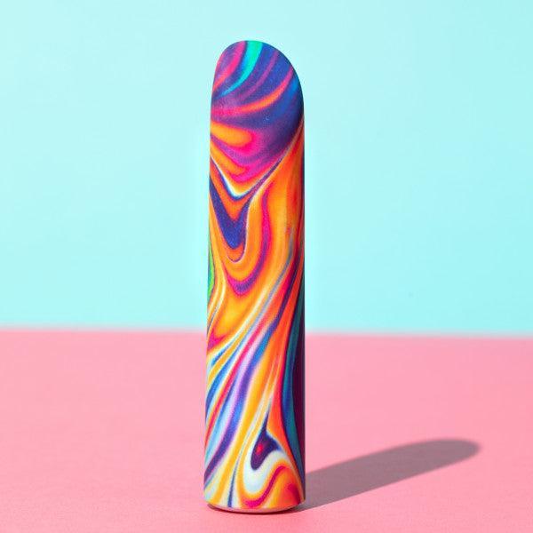 The Limited Addiction Power Bullet Vibe - Psyche by Blush is a colorful, cylindrical device with a marble pattern featuring swirls of vibrant pink, orange, yellow, blue, and purple hues. It stands upright on a pink surface against a light blue background. This USB rechargeable vibe boasts 10 different vibrations and casts a shadow to the right.