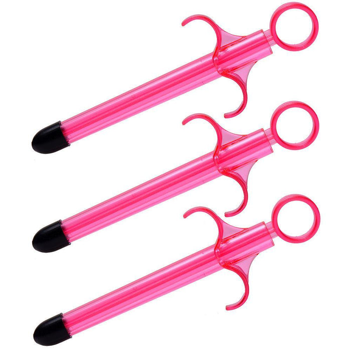 Three identical pink plastic syringes with ring pull handles and black tips from the Lubricant Launcher 3-Pack by XR Brands are arranged diagonally across the image. The playful design, reminiscent of medical syringes, is clearly intended for non-serious use, perhaps as a Lubricant Launcher or for vaginal moisturizing.