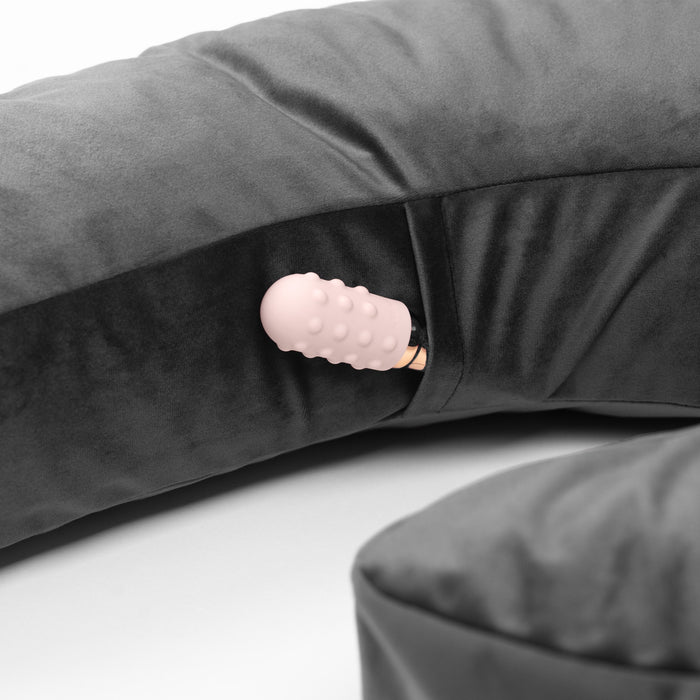A close-up of the Liberator Lune Snuggle Pillow with Vibrator Pocket in dark gray, showing a pink textured object partially visible in its small pocket. Its plush fabric and smart design add comfort and convenience.