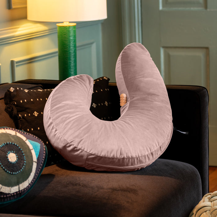 The Liberator Lune Snuggle Pillow with Vibrator Pocket, in light pink and U-shaped, sits on a dark sofa beside a black-and-white patterned cushion. A round decorative pillow and a green lamp appear in the background.