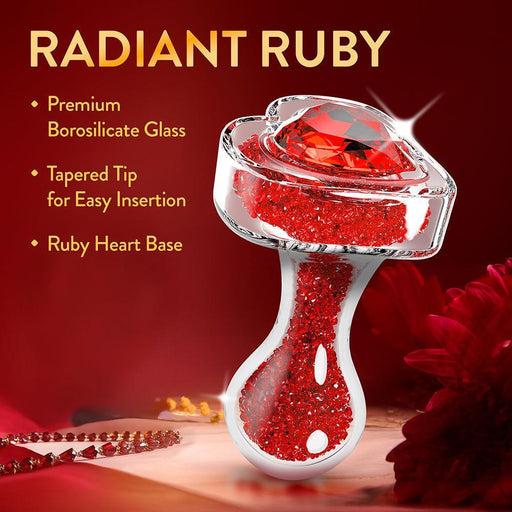 The Lustre Glass Butt Plug with Gem Base & Red Crystals features premium borosilicate glass, a tapered tip for easy insertion, and a stunning heart-shaped ruby gem base sparkling with red crystals. Displayed on a red floral background.