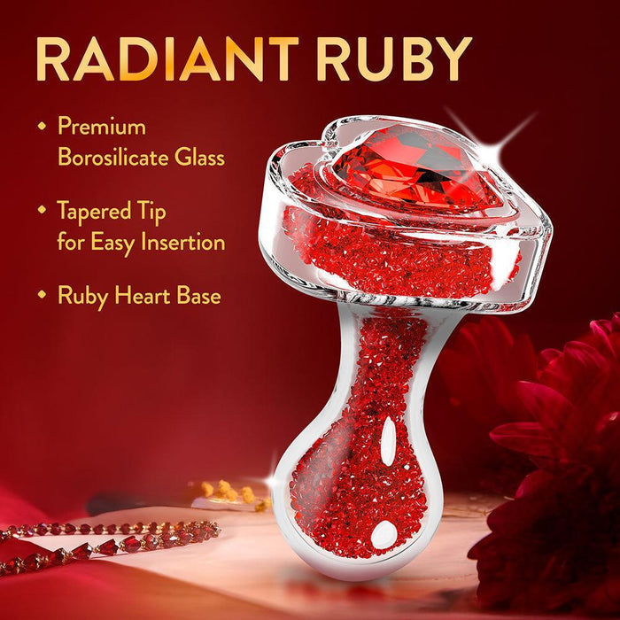 The Lustre Glass Butt Plug with Gem Base & Red Crystals features premium borosilicate glass, a tapered tip for easy insertion, and a stunning heart-shaped ruby gem base sparkling with red crystals. Displayed on a red floral background.