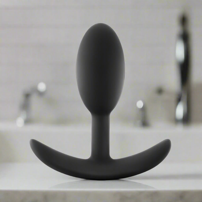 The Luxe Small Wearable Silicone Vibra Slim Plug in black, by Blush, features a smooth, oval design with a curved base for personal use. Made of hypoallergenic body-safe silicone, this plug is soft and flexible to ensure comfort and ease of insertion.