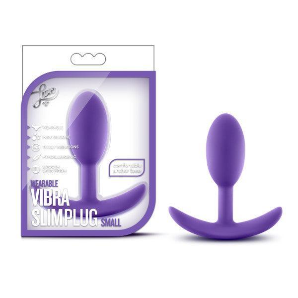 A small, purple silicone anal plug with a comfortable anchor base displayed in front of its packaging. The Luxe Small Wearable Silicone Vibra Slim Plug by Blush ensures safe use with its hypoallergenic material. The packaging features product details and a clear window showcasing the plug, labeled as "Wearable Vibra Slim Plug Small.