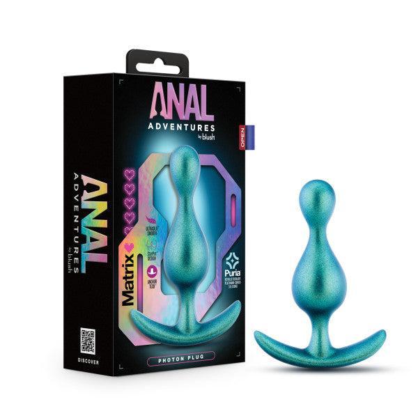 A shimmery green anal plug, crafted from platinum-cured silicone, sits beside its packaging. The box features the brand "Blush" and a label indicating "Matrix First Time Soft Silicone Butt Plug." The package has a holographic design with additional information about the product and specific details for beginners.
