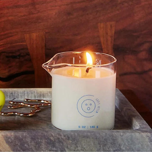 A lit Melt Together Massage Oil Candle by Dame (Green Tea, Fig & Cedarwood) in a clear beaker with blue markings and the Wild Lust logo sits on a gray tray, with gold décor and a green ball partially visible beside it.