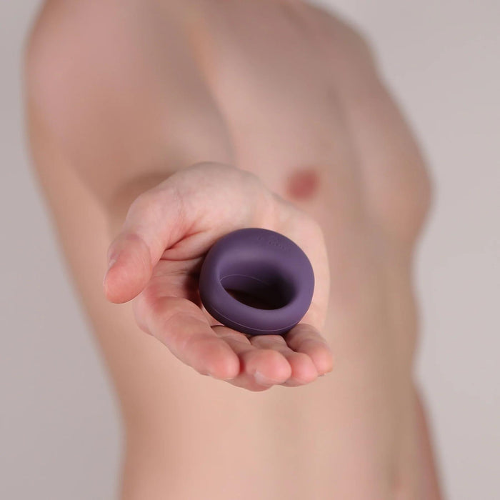 A shirtless person holds the Midnight Magic Couples' Sex Toy Gift Set's small, purple vibrating ring in their outstretched hand against a neutral background.
