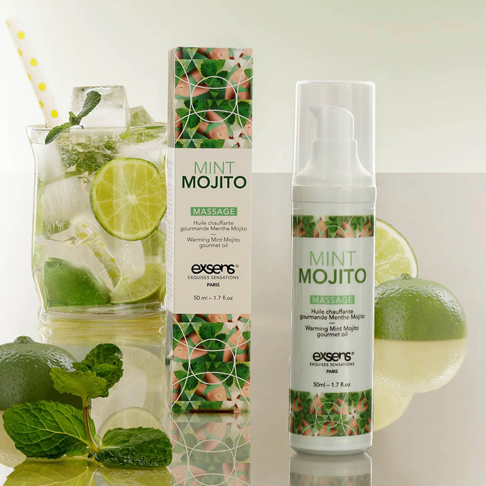 A bottle and box of Exsens Mint Mojito Flavored Warming Massage Oil 50ml are displayed beside a mojito glass with lime, mint, and ice on a reflective surface with fresh mint and lime. Enjoy this vegan massage oil for a refreshing touch.