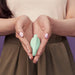 A person with light pink nails holds the Elvie Trainer Smart Kegel Exerciser (mint green) in cupped hands against a purple background, wearing a beige top and olive-green skirt.