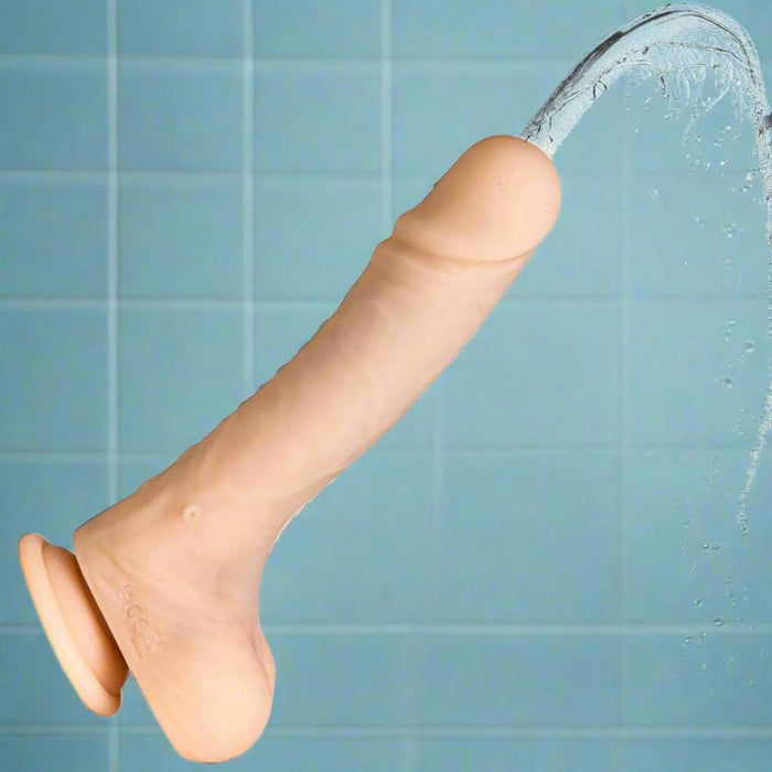 The Money Shot 8 Inch Vibrating Squirting Silicone Dildo (Ejaculating Strap on) in Vanilla is shown with its suction base attached to a tiled shower wall, squirting water from its tip.